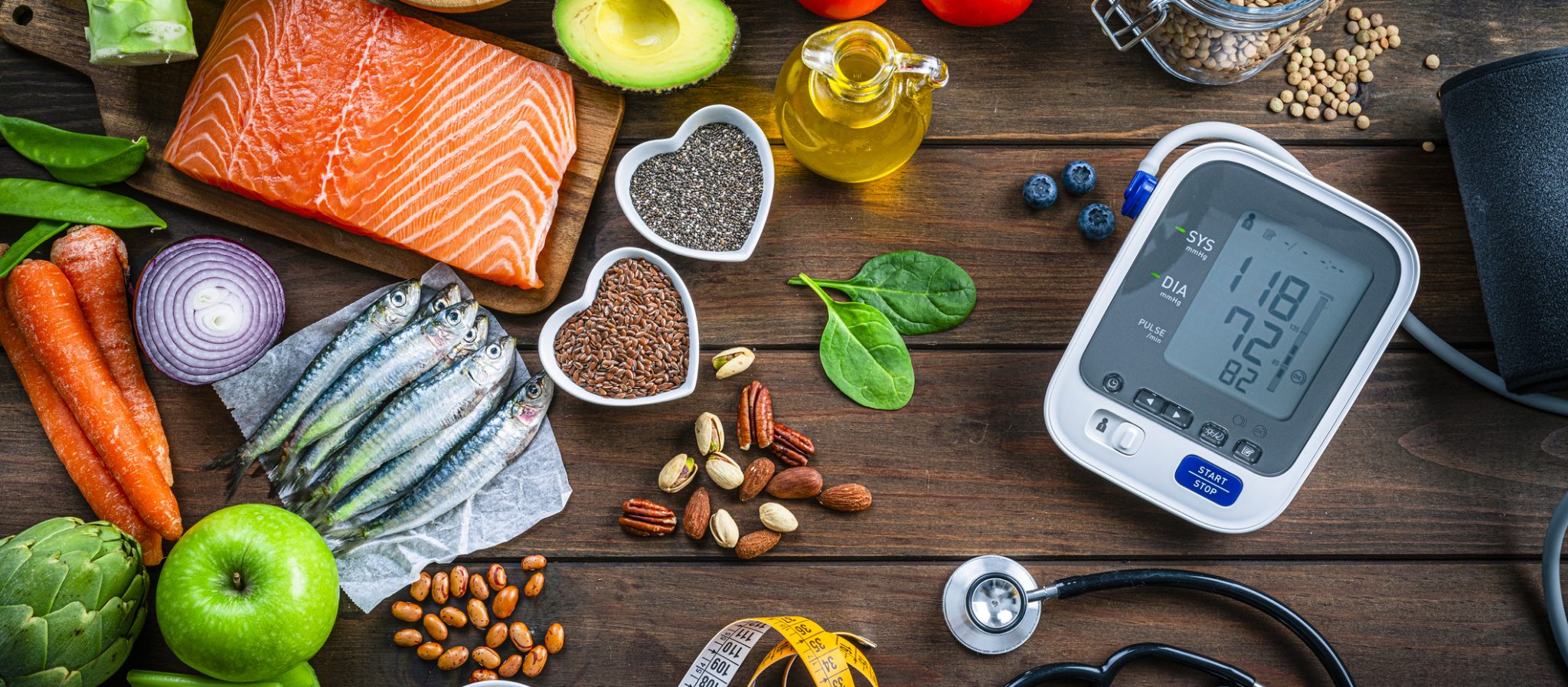 Healthy food for heart care: overhead view of healthy food rich in Omega-3 and antioxidants and a blood pressure monitor, tape measure and stethoscope shot on wooden table. The composition includes a salmon fillet, sardines, avocado, extra virgin olive oil, brown lentils, celery, artichoke blueberries, celery, carrots, brown lentils, asparagus, artichoke, broccoli, flax seeds, chia seeds and some nuts like almonds, pistachio and pecan. High resolution 42Mp studio digital capture taken with SONY A7rII and Zeiss Batis 40mm F2.0 CF lens